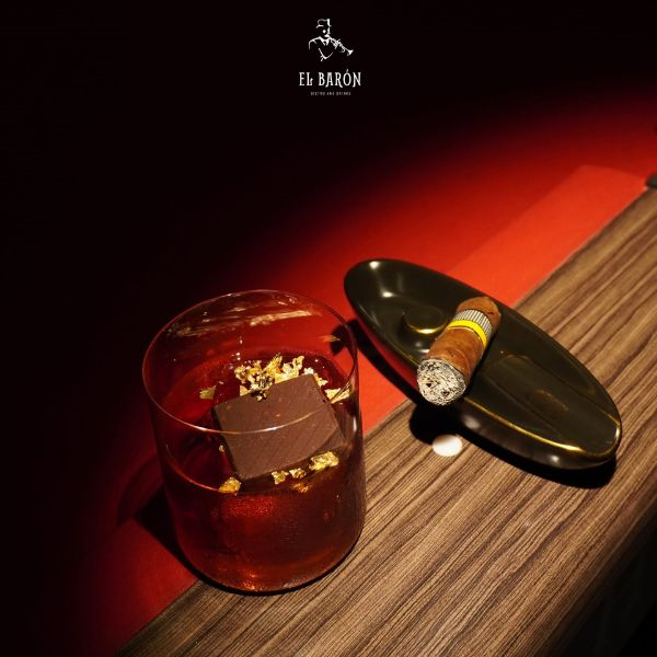 Gold-flake garnished cocktail glass paired with a premium lit cigar resting on a ceramic ashtray at El Baron Whiskey and Cigar Bar